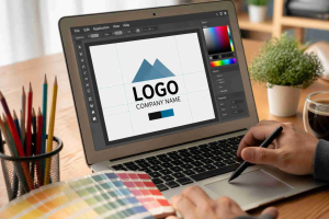 Desain logo dengan tulisan 'LOGO' di layar laptop, dengan latar belakang pensil warna dan palet warna, menggambarkan proses kreatif di ruang kerja.